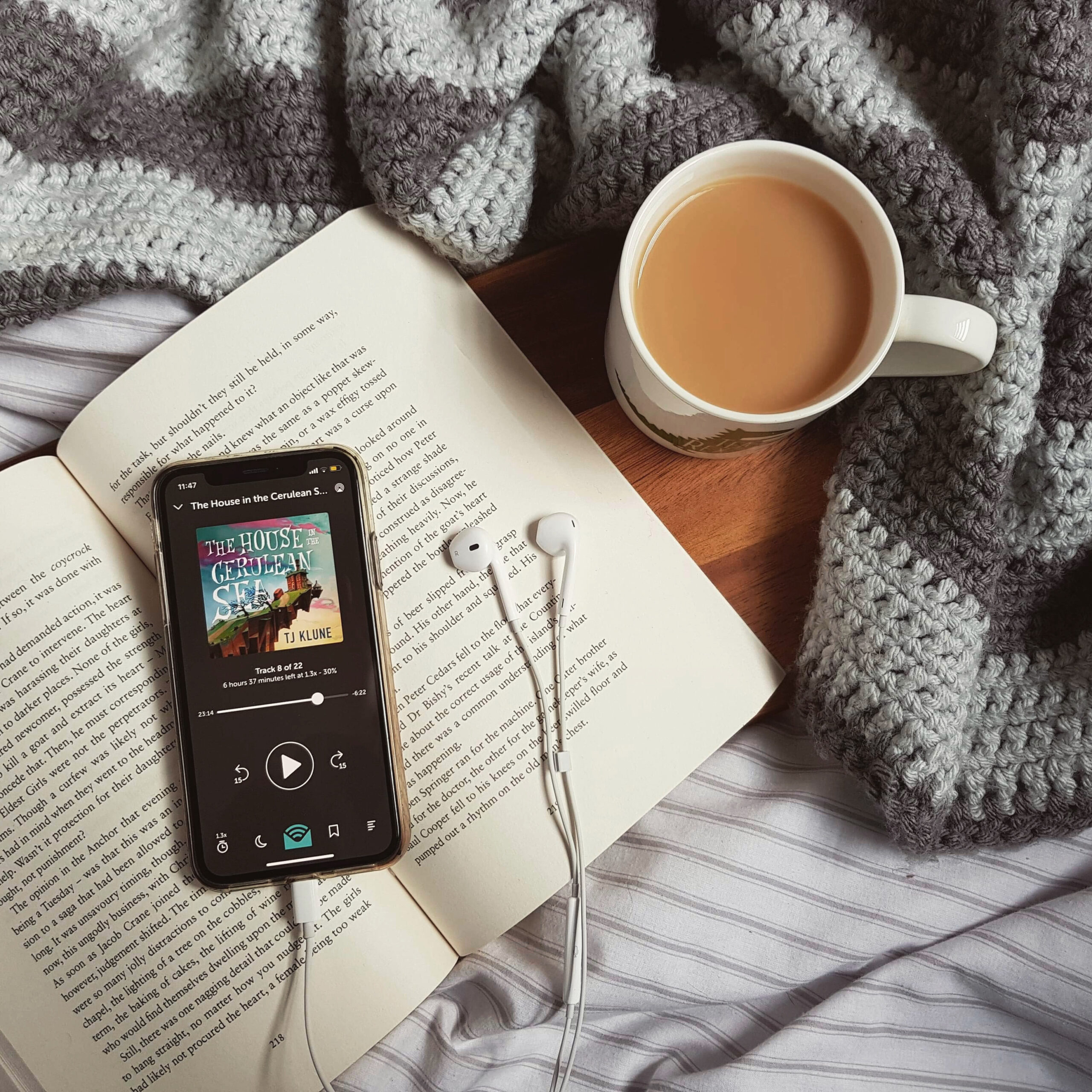 An iPhone lying on an open book shows an audiobook of The House in the Cerulean Sea by T. J. Klune. There are earphones next to the book, as well as a cup of tea on a brown wooden board, surrounded by a dark green velvet blanket.
