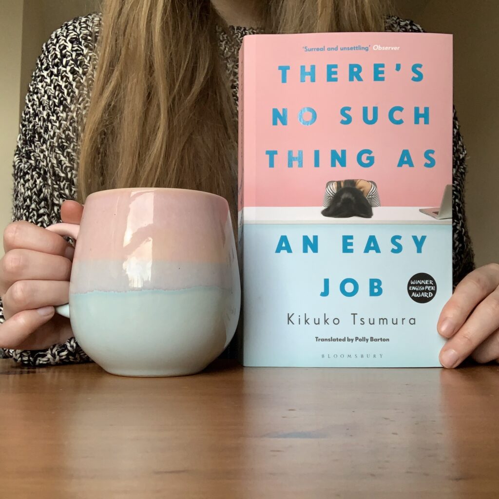 Abbie sits at a brown desk, holding  a copy of There’s No Such Thing As an Easy Job by Kikuko Tsumura, tr. Polly Barton* and a pink and blue cup of tea.