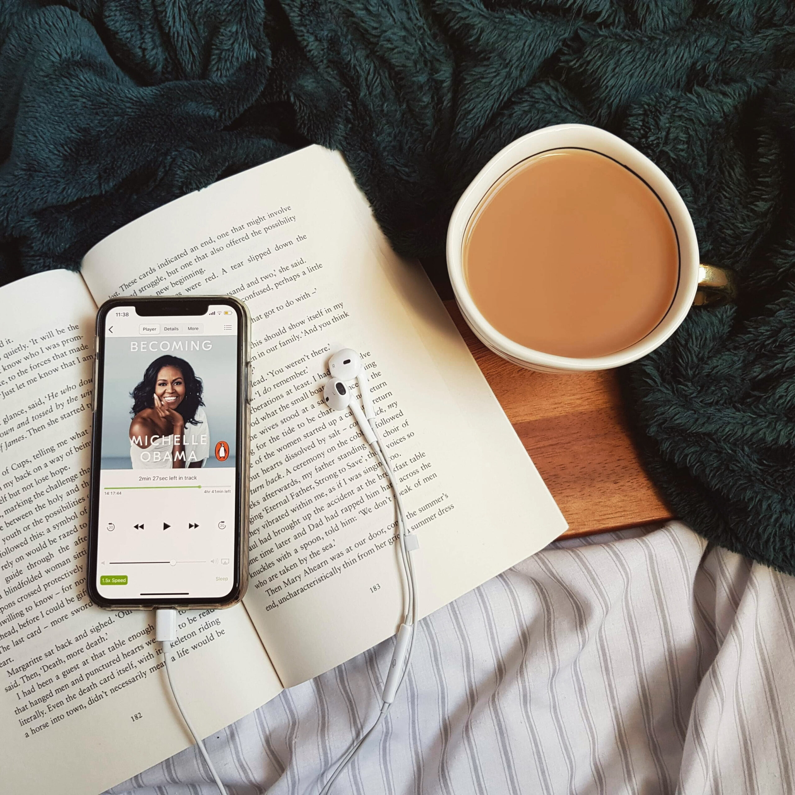 An iPhone lying on an open book shows an audiobook of Becoming by Michelle Obama. There are earphones next to the book, as well as a cup of tea on a brown wooden board, surrounded by a dark green velvet blanket.
