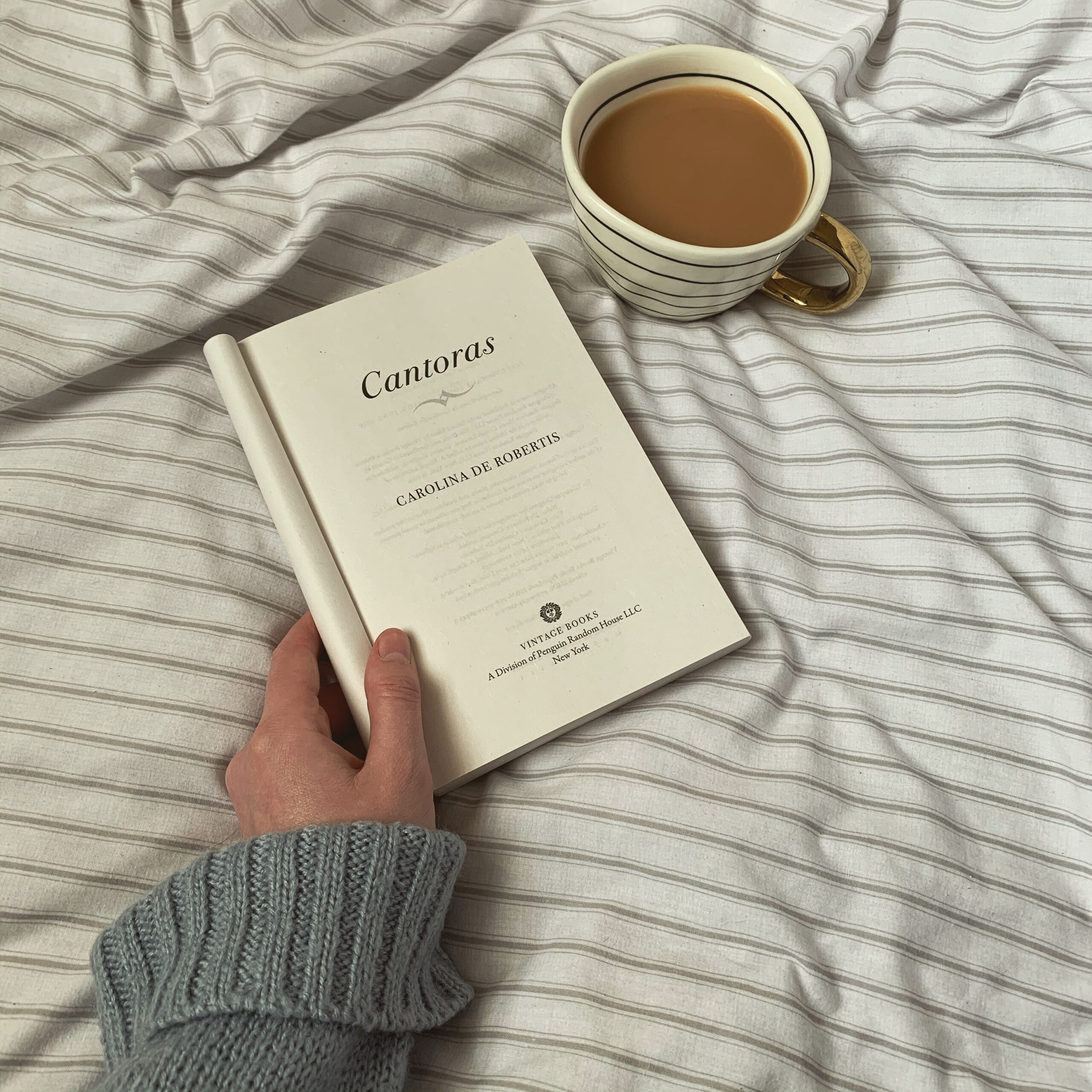 A paperback of Cantoras by Carolina de Robertis sits open to the title page on a striped bedspread, with a cup of tea to the top right.