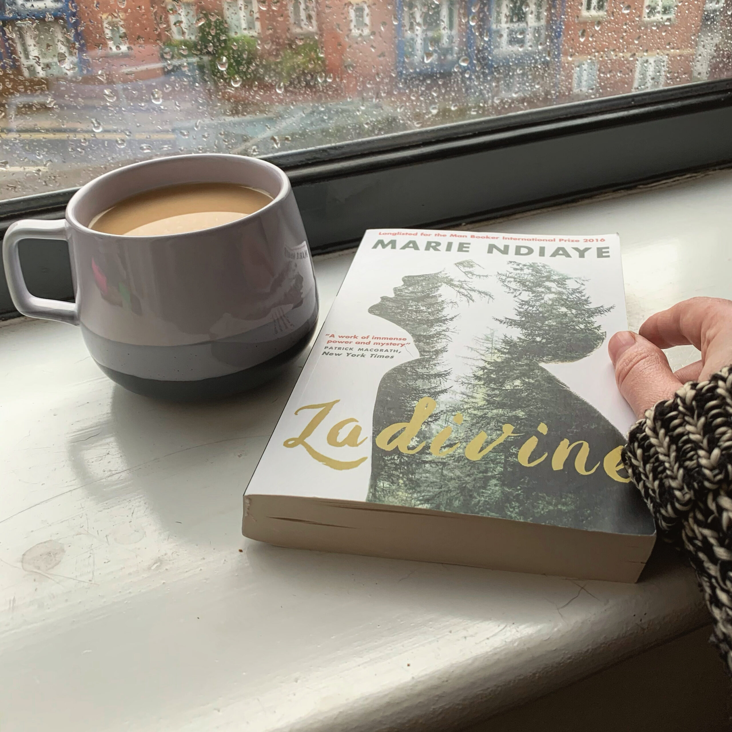 A copy of Ladivine by Marie NDiaye, tr. Jordan Stump sits on a white windowsill while a purple cup of tea sits next to it. 