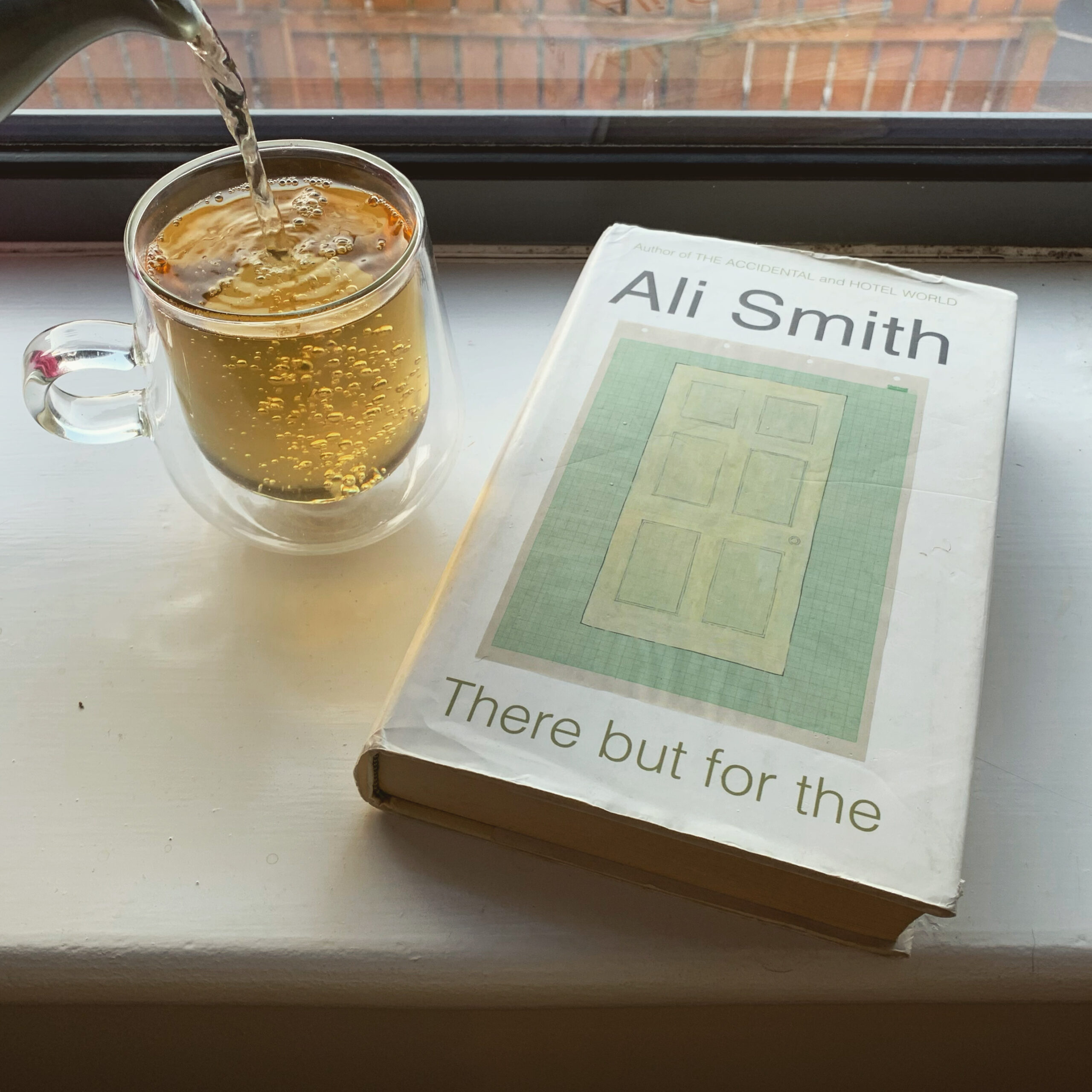 A copy of There But For The by Ali Smith lies on a white windowsill, while next to it tea is being poured into a clear glass cup. 